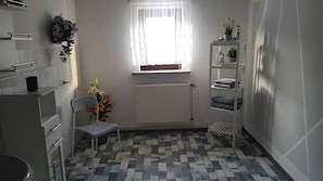 A small, modern bathroom with a tiled floor, a white radiator, a window with sheer curtains, a chair, a shelf with towels, and a potted plant.