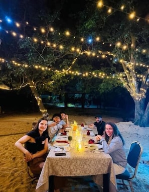 Un área de comedor al aire libre con una mesa lista para una comida, rodeada de árboles y luces colgantes.