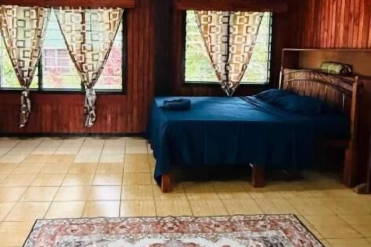 Una habitación con cama, dos ventanas con cortinas, un tapete estampado y paredes de madera.