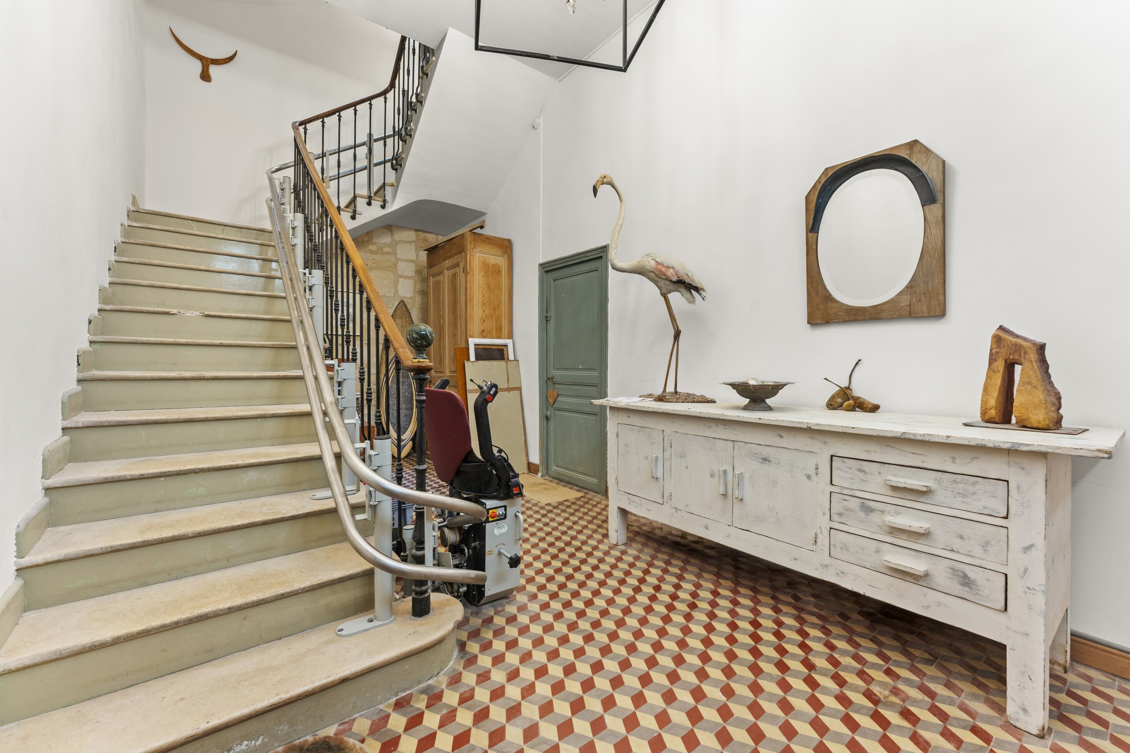 Una escalera con barandilla metálica, una puerta de madera, un armario blanco con cajones, un espejo y una estatua de flamenco.
