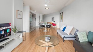 A modern living room with a glass coffee table, a sofa, a TV, and a dining area with a red chair. - Modern Inner-City Stay in Valley|Heart of Brisbane (Fortitude Valley)