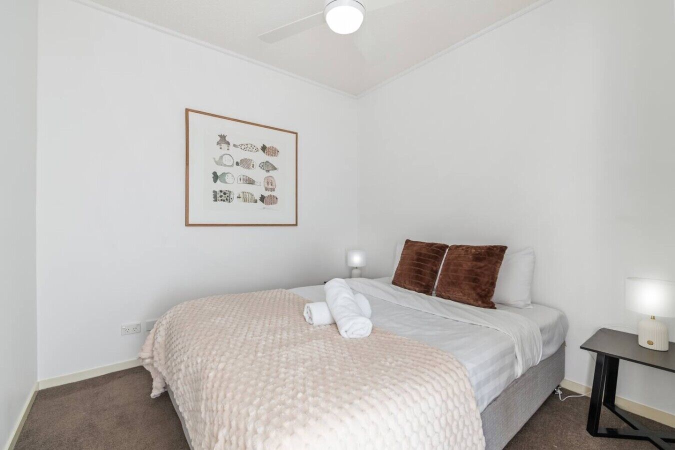 Un dormitorio con cama, almohadas, una mesita de noche, lámpara, ventilador de techo y un cuadro en la pared.
