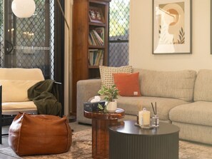 A living room with a sofa, a coffee table, a bookshelf, and a window with a view. - Stylish Family Home w/ A/C, 5 mins to Westfield (Upper Mount Gravatt)