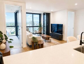 A modern living room with a large TV, a dining area, and a view of the city. - 2BR Story Bridge Views | Resort Pool & Parking (Brisbane)