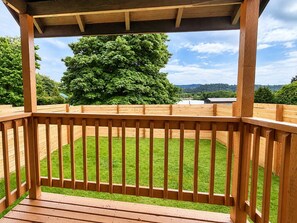 Una terraza de madera con barandilla, un jardín cubierto de césped y un gran árbol al fondo.