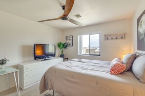 Ein Schlafzimmer mit einem Bett, einem Deckenventilator, einem Fernseher, einem Fenster, einer Pflanze und einem an der Wand befestigten Regal.