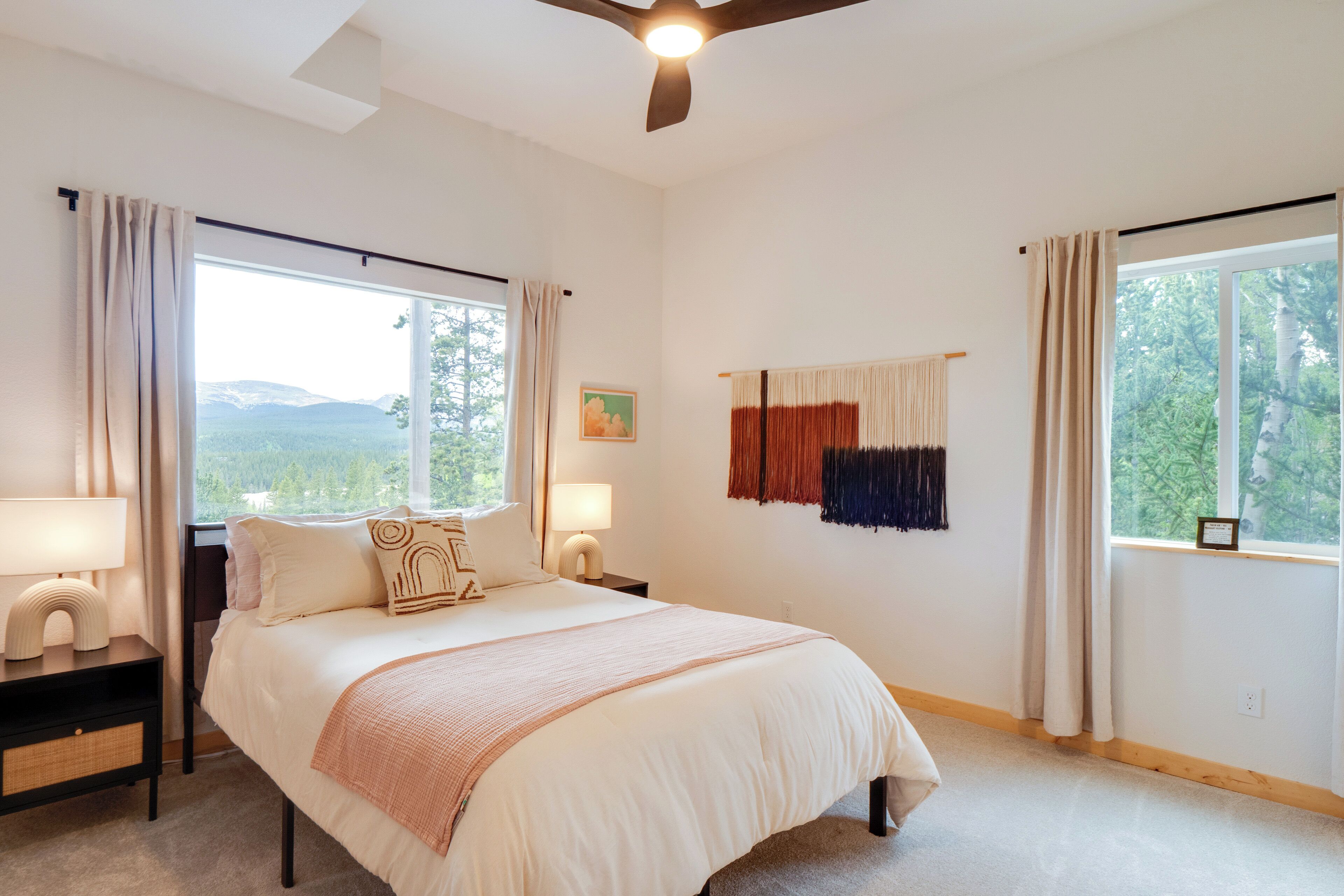 A bedroom with a large bed, two bedside tables with lamps, a ceiling fan, and a view of the outdoors through a window.
