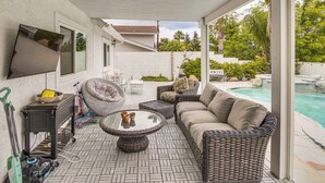Une terrasse couverte avec des meubles en osier, une piscine et un téléviseur fixé au mur.