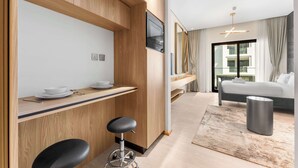 Ett modernt hotellrum med ett litet kök i trä, en säng, ett stort fönster och en minimalistisk inredning.