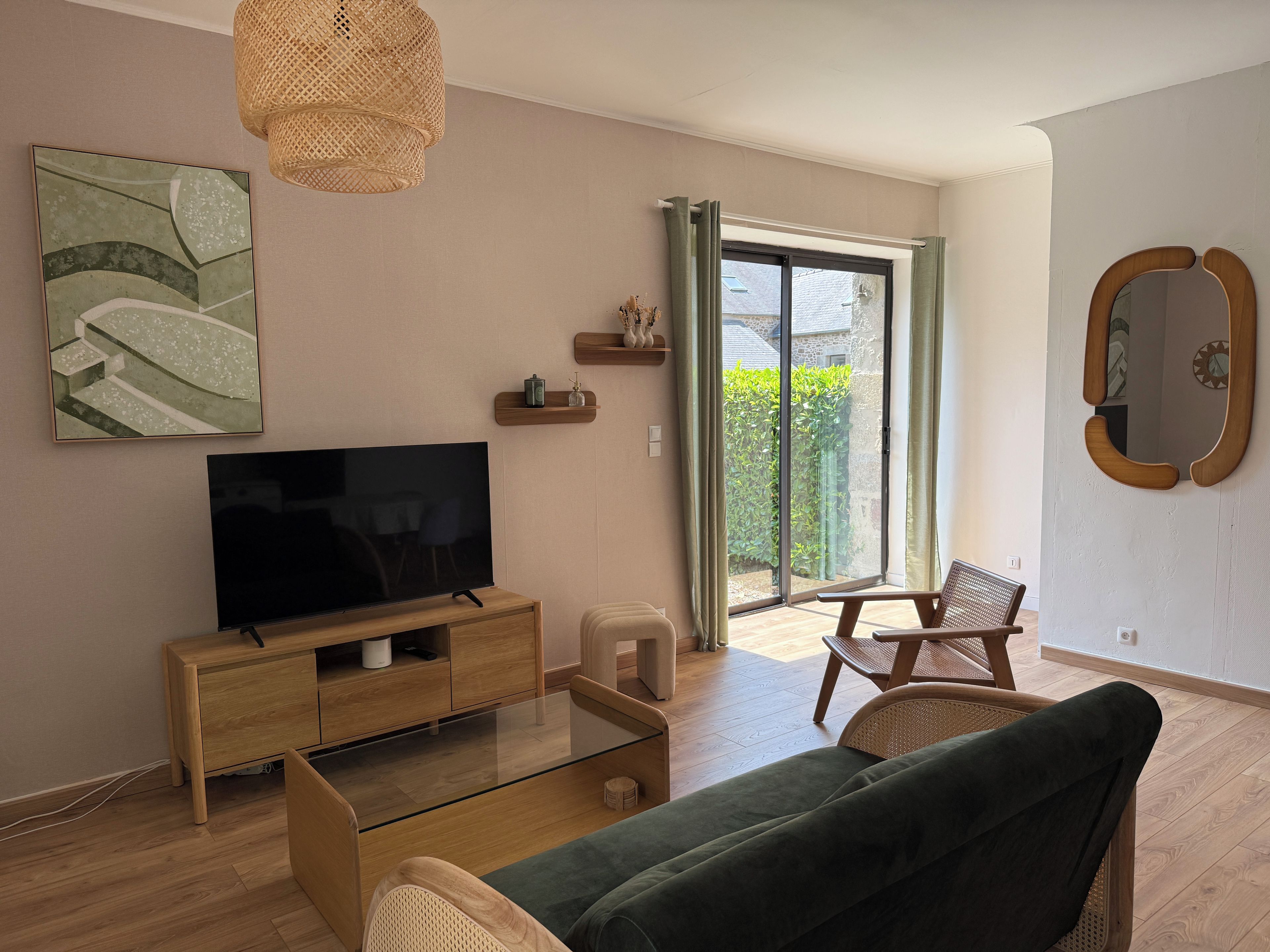 A living room with a flat-screen TV on a wooden stand, a green sofa, a wicker chair, and a large window with curtains.