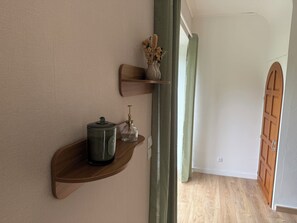 A room with a wooden shelf holding a kettle and a glass container, a wooden door, and a green curtain. - Maison Saule | A Charming Retreat on the Emerald Coast (Val-d'Arguenon)