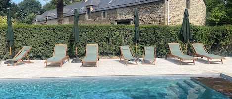 Un área junto a la piscina con sillones de descanso y sombrillas, un edificio de piedra al fondo y una piscina azul cristalina.