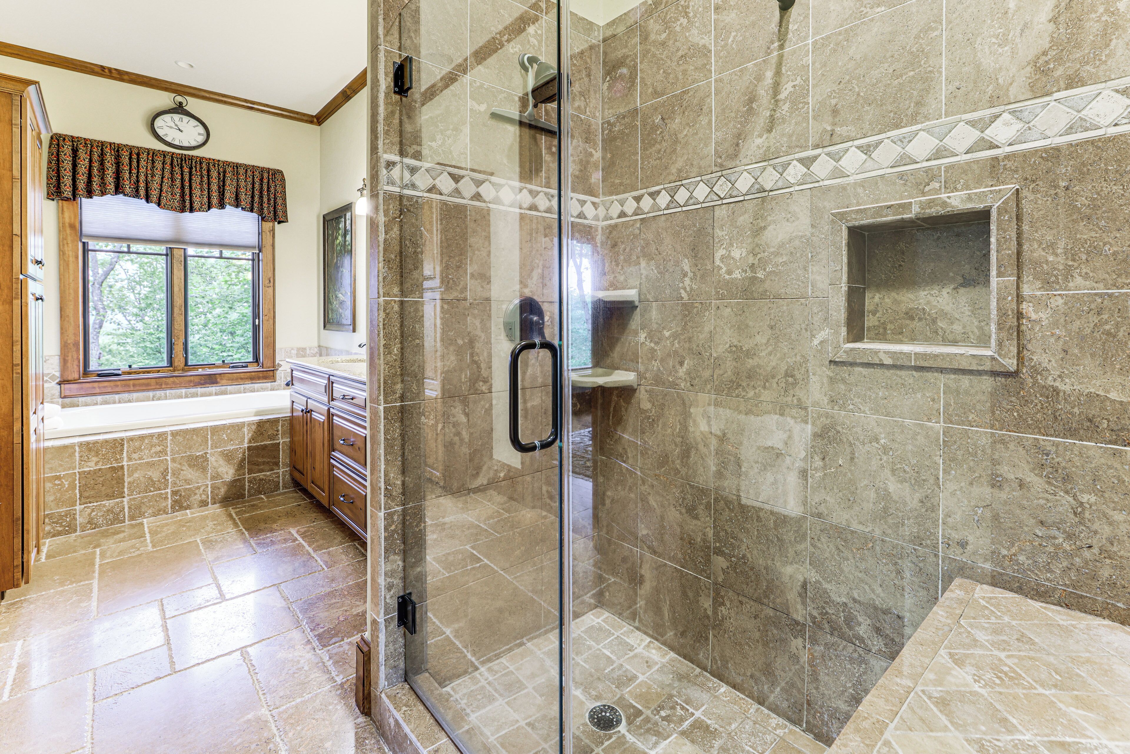 A bathroom with a glass shower enclosure, a window with a patterned valance, and a tiled floor.
