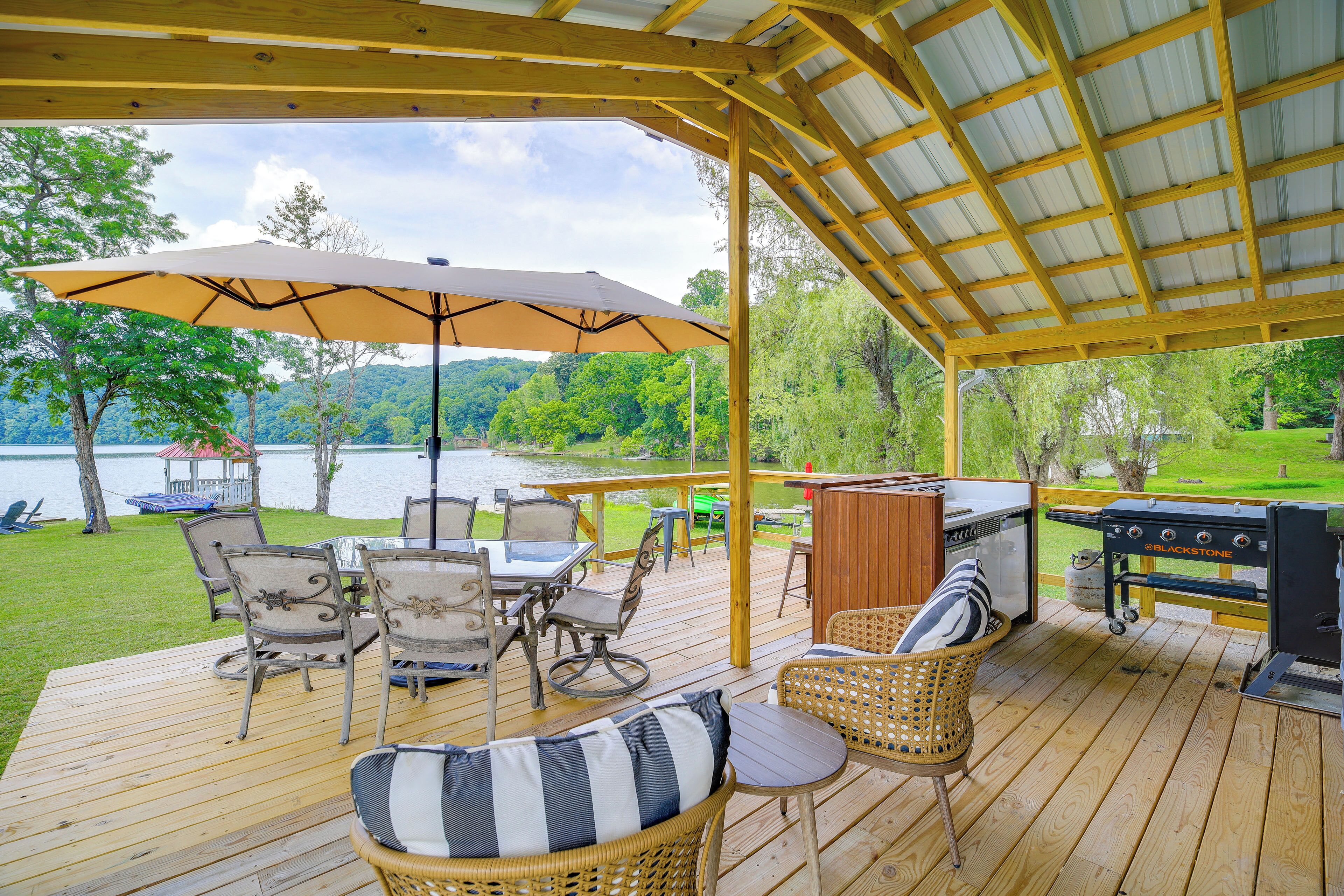 Una terraza techada con mobiliario exterior, una parrilla y vistas a un lago.