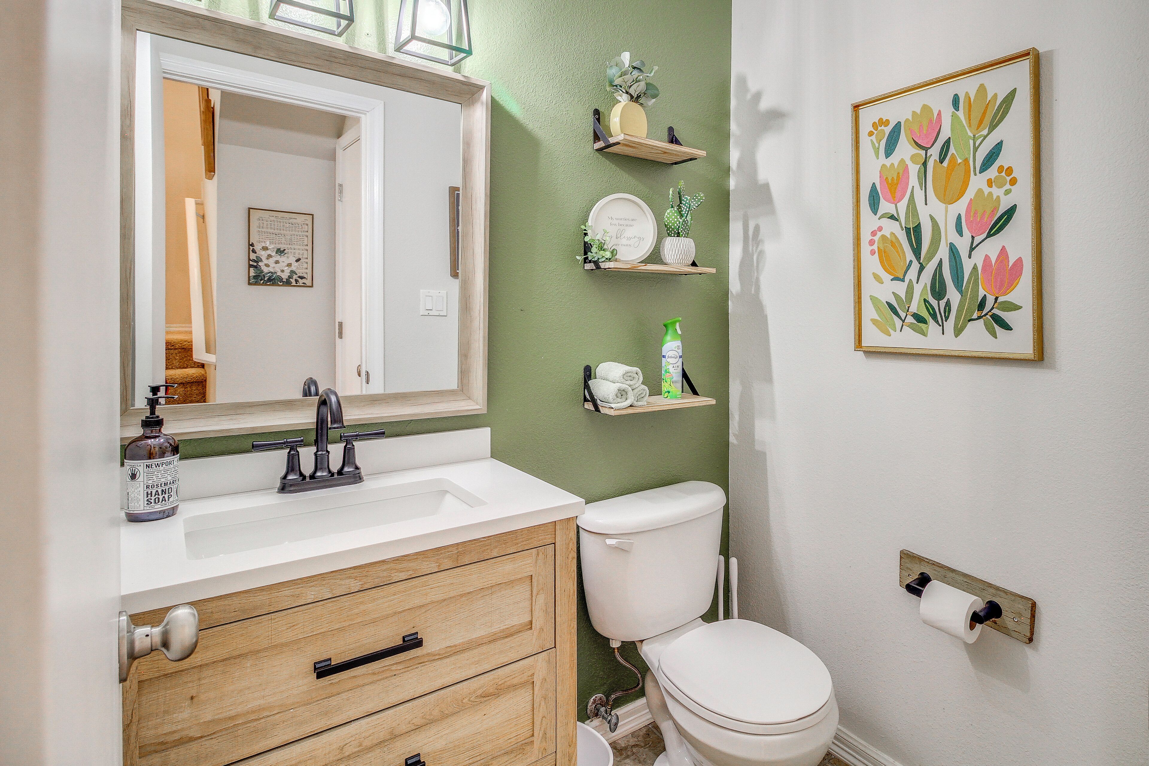 Un baño con lavamanos blanco, tocador de madera, inodoro y estantes decorativos con plantas y una obra de arte floral enmarcada.