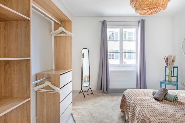 Ein Schlafzimmer mit einem Holzschrank, einem Spiegel, einem Bett mit beiger Decke und einem Fenster mit Vorhängen.