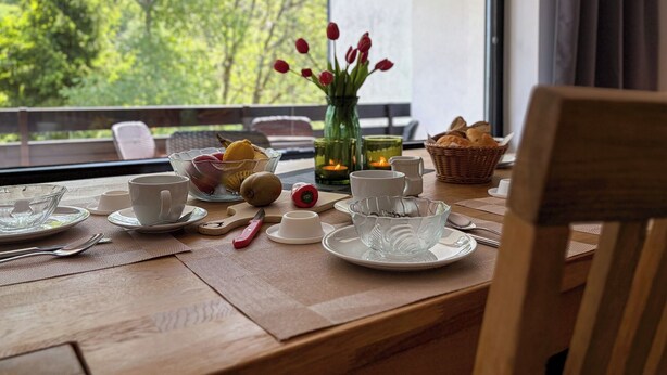 Una mesa lista para comer, con platos, tazas, un frutero y un jarrón con flores rojas.