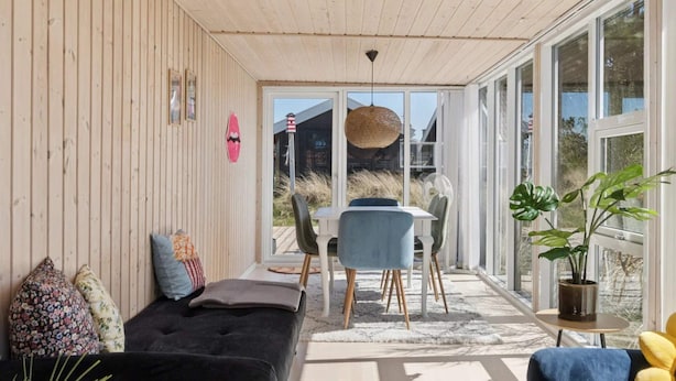 Ein gemütliches Zimmer mit Holzbalkendecke, einem schwarzen Sofa, einem Esstisch mit Stühlen und großen Fenstern.