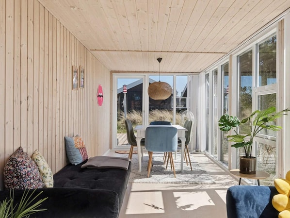 Ein gemütliches Zimmer mit Holzbalkendecke, einem schwarzen Sofa, einem Esstisch mit Stühlen und großen Fenstern.