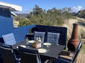 An outdoor dining area with a table set for two, surrounded by wicker chairs with blue cushions, a blue fence, and a rustic chimney. - 4 Star Holiday Home in Thisted (Thisted)