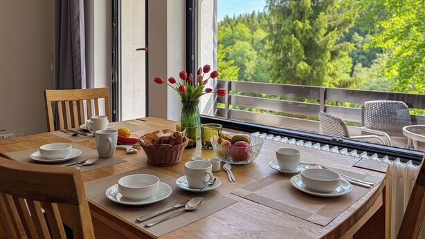 Ein Holztisch ist mit Tellern, Tassen und einem Brotkorb gedeckt, mit Blick auf einen Balkon und eine grüne Aussicht.