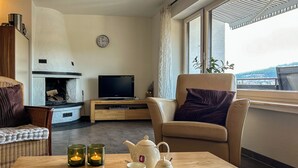 A living room with a fireplace, a television, a sofa, a chair, a table with a book, a teapot, and candles. - Cozy vacation apartment with a view, starting point for hikes, near Winterberg (Winterberg)