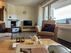 A living room with a fireplace, a television, a sofa, a chair, a table with a book, a teapot, and candles. - Cozy vacation apartment with a view, starting point for hikes, near Winterberg (Winterberg)