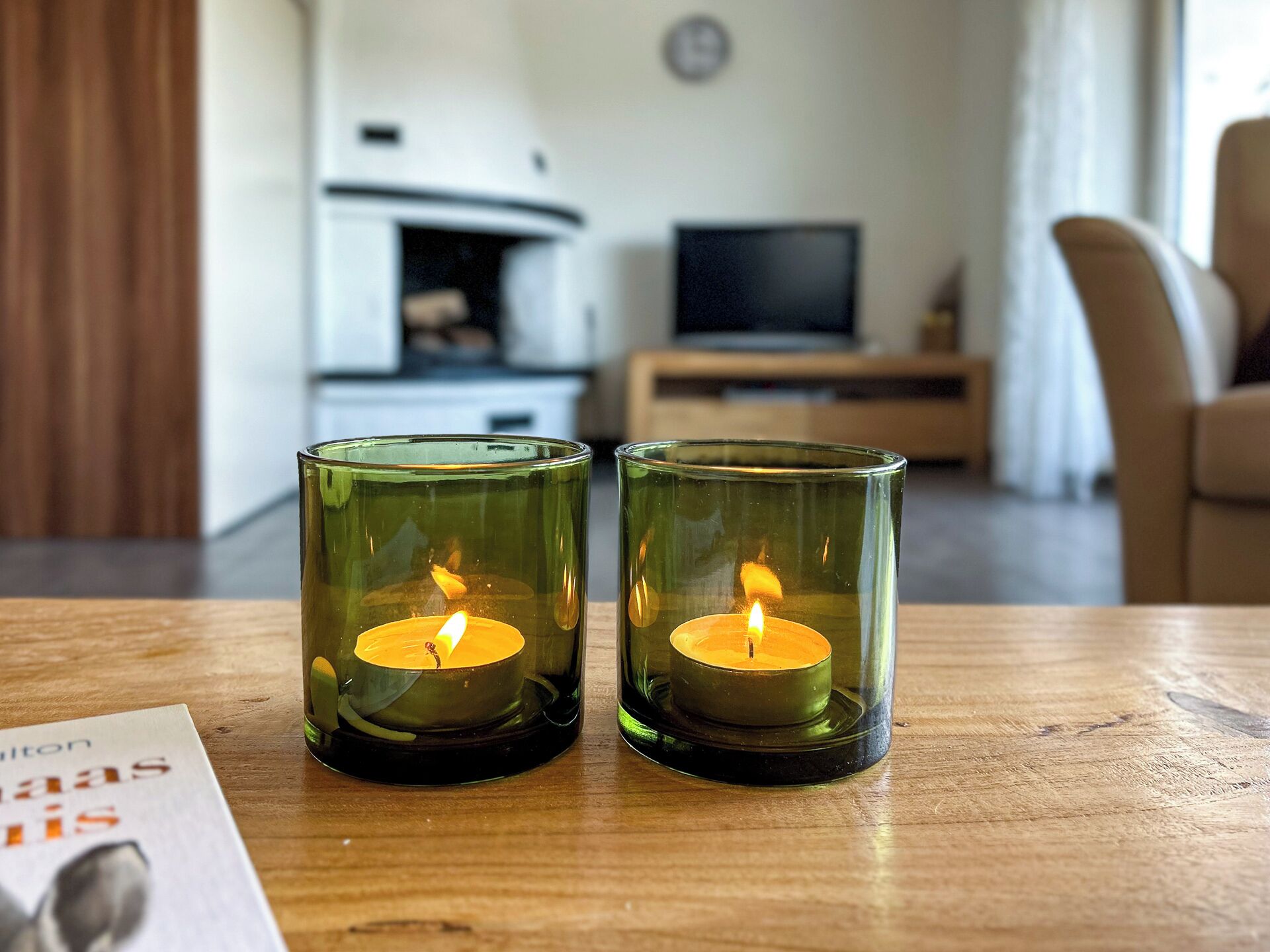 Two lit candles in glass holders on a wooden table in a living room with a TV and a sofa.