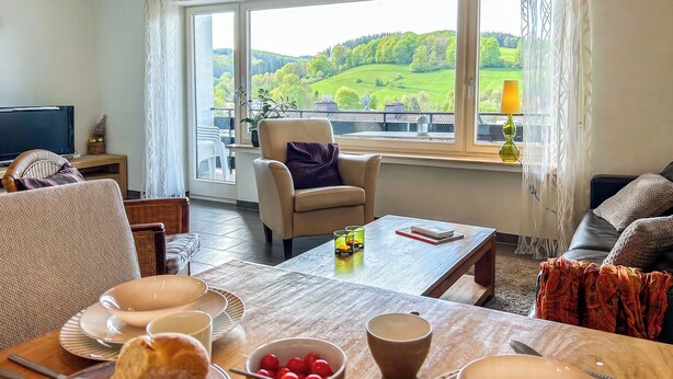 A living room with a large window, a sofa, a chair, a coffee table, and a dining table with plates, cups, and a bowl of cherries. - Cozy vacation apartment with a view, starting point for hikes, near Winterberg (Winterberg)