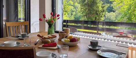 A dining table set with plates, bowls, cutlery, and a vase of tulips, with a view of trees through a balcony railing.