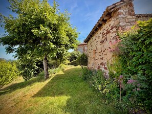 Um edifício rústico de pedra com telhado inclinado, rodeado por vegetação exuberante e um céu claro.