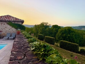 Um edifício de pedra com telhado de telhas, piscina e cadeiras de descanso.