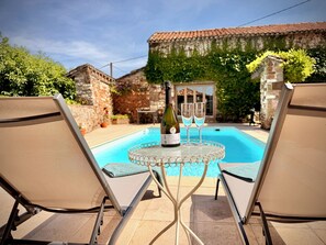 Área da piscina com espreguiçadeiras, uma mesa com garrafa e copos, e um edifício de pedra ao fundo.