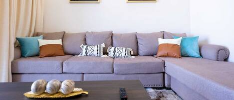 Una sala de estar con un sofá seccional gris, varios cojines, una mesa de centro con objetos decorativos y dos cuadros en la pared.