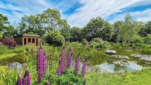 En have med et bassin, lilla blomster og en trækonstruktion.