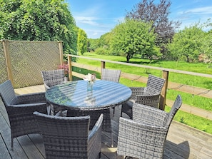 Ein runder Glastisch mit Korbstühlen auf einer Holzterrasse, mit Blick auf einen grünen Garten mit Bäumen.
