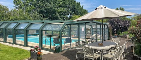 Een overdekte zwembad met een terras, buitenmeubels en een grote parasol.