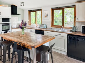 Una cocina moderna con una mesa de madera, taburetes de bar, lavavajillas, fregadero y ventana con vistas a la vegetación.