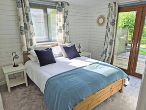 A bedroom with a bed, bedside table, lamp, and a view of the outdoors. - Torridge Lodge - UKC9517 (Sheepwash)