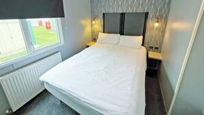 A bedroom with a bed, a window with a view, a radiator, and two bedside lamps. - Palm Grove 1 - UKC9556 (Chapel St Leonards)