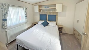 Une chambre à coucher avec un lit, une fenêtre avec des rideaux, un radiateur, une tête de lit en bois, une étagère avec des objets décoratifs et une armoire.