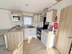 A compact kitchenette with a microwave, stove, sink, and storage cabinets. - Palm Grove 182 - UKC9560 (Chapel St Leonards)