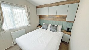 A bedroom with a bed, a nightstand, a lamp, a radiator, and a window with curtains. - Skegness Fields 108 - UKC9566 (Skegness)