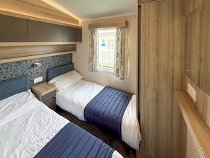 A bedroom with two beds, a window with curtains, a headboard with a geometric pattern, and a wooden wardrobe. - Sunnymede A6 - UKC9568 (Ingoldmells)
