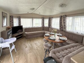Interior moderno de una casa rodante, con zona de comedor, asientos, televisor y pequeña cocina.