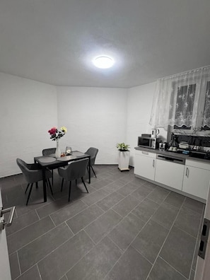 Eine kleine Küche mit Esstisch, Stühlen, einer Kaffeemaschine und einem Blumentopf.