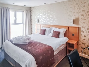 A hotel room with a bed, a nightstand, a chair, a window with curtains, and a wall with a leaf pattern.