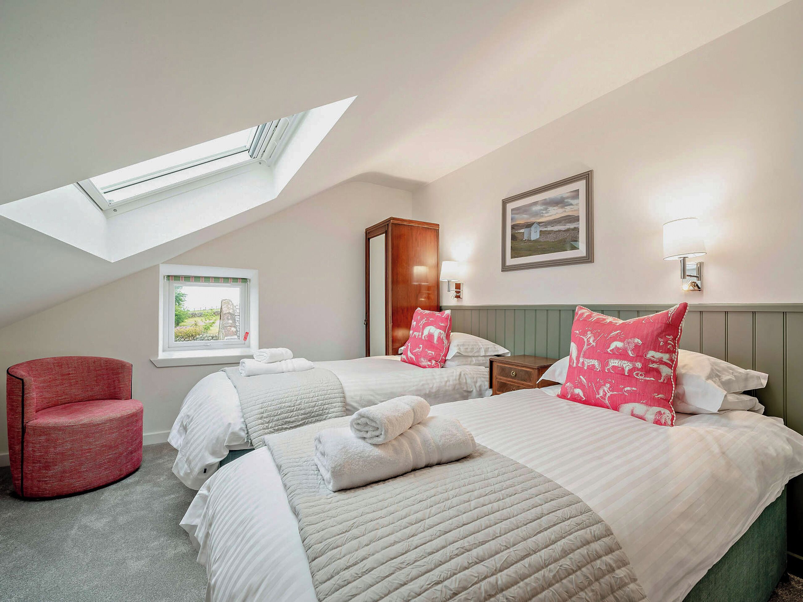 A hotel room with two beds, a skylight, a red chair, a window with a view, a framed picture, and a wall-mounted lamp.