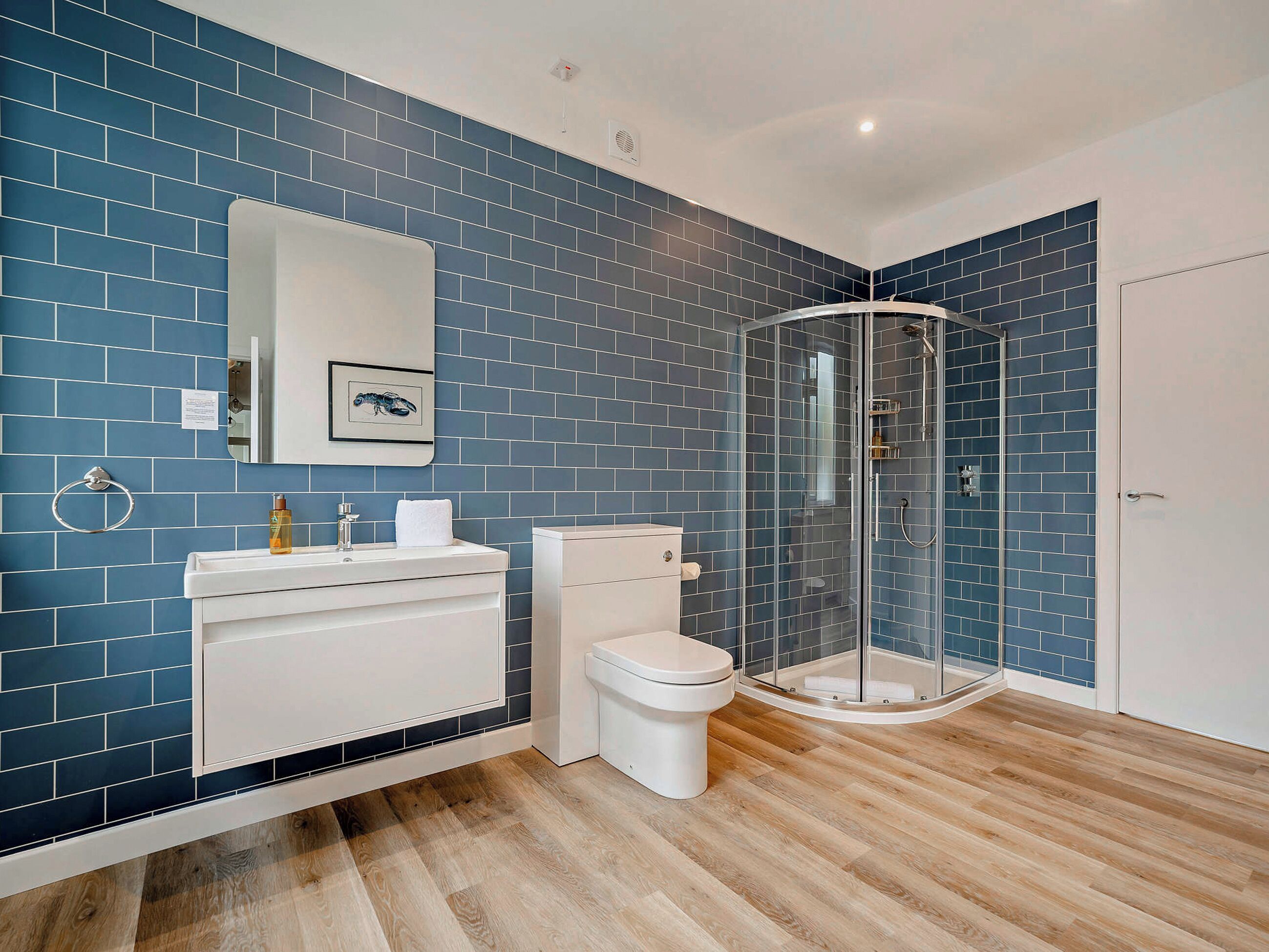 Une salle de bain avec des murs recouverts de carreaux bleus, un lavabo blanc, des toilettes, une douche en verre et un plancher en bois.