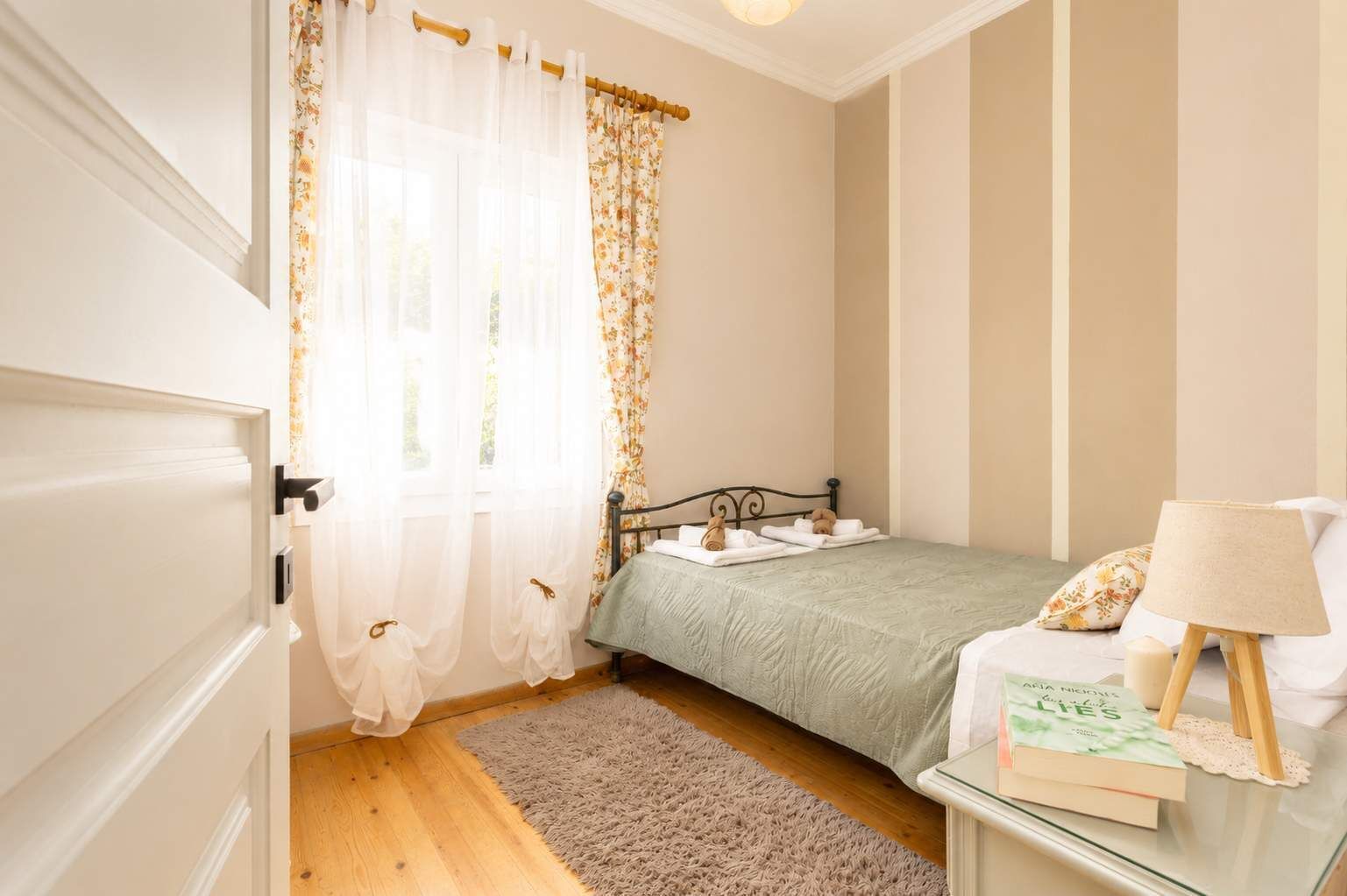 Un dormitorio con una cama, una mesita de noche con lámpara, un libro y una ventana con cortinas traslúcidas.